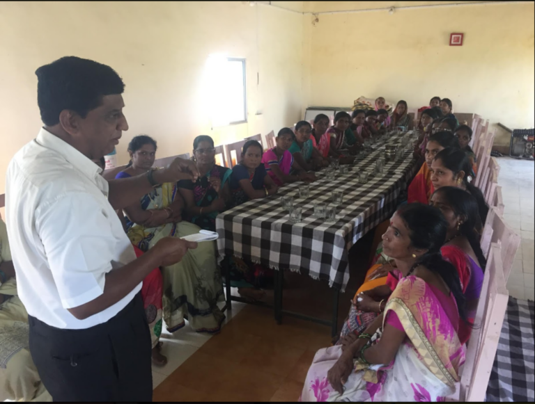 At a long table, a group of people sit and listen to a speaker who stands before them.